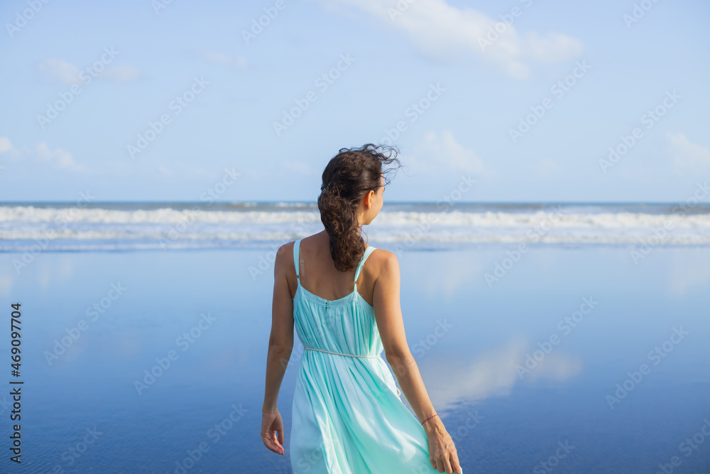 Back view portrait of beautiful Caucasian woman on the beach. Summer vacation in Asia. Young woman wearing dress. Blue sky. Ocean with horizon line. Travel lifestyle. Bali Back view portrait of beautiful Caucasian woman on the beach. Summer vacation in Asia. Young woman wearing dress. Blue sky. Ocean with horizon line. Travel lifestyle. Bali