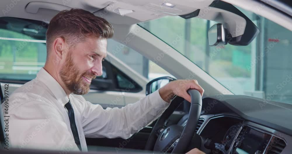 Caucasian bearded man showing thumb up and smiling on camera while sitting in side new car at modern salon. Concept of people, transport and purchase. Caucasian bearded man showing thumb up and smiling on camera while sitting in side new car at modern salon. Concept of people, transport and purchase.