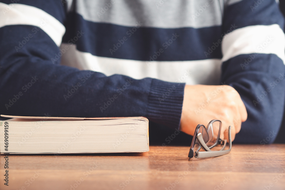 Education, knowledge, and relaxation concept. A man in a sweater with a book and glasses on a wooden table while sitting in the library. Close-up photo