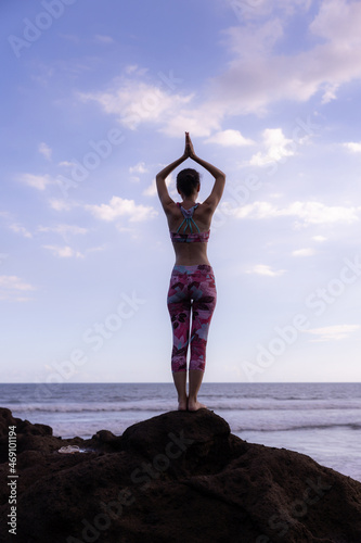 Woman standing on the rock, practicing yoga. Young woman raising arms with namaste mudra at the beach. Blue sky background. Yoga retreat in Bali. View from back. Copy space.
