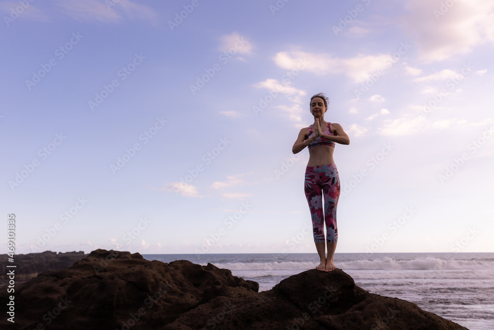 Slim woman standing on the rock, practicing yoga at the beach. Hands in namaste mudra. Blue sky background. Yoga retreat. Concentration and balance. Horizontal layout. Copy space. Bali