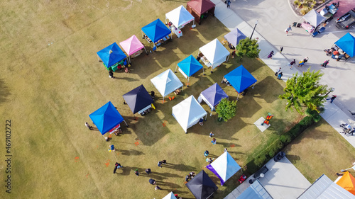 Top view busy local people on sidewalk and grassy lawn shopping in colorful vendor tents at farmer market near Dallas, Texas, USA