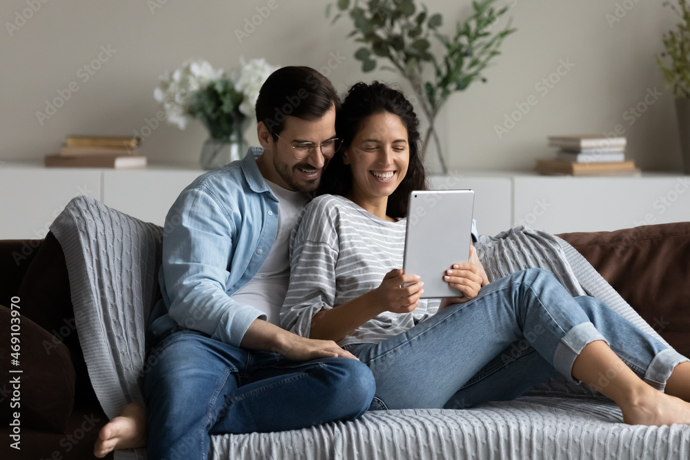 Happy couple hugging, using tablet together, relaxing on couch, smiling beautiful woman and man in glasses looking at screen, chatting online by video call, shopping or watching movie at home