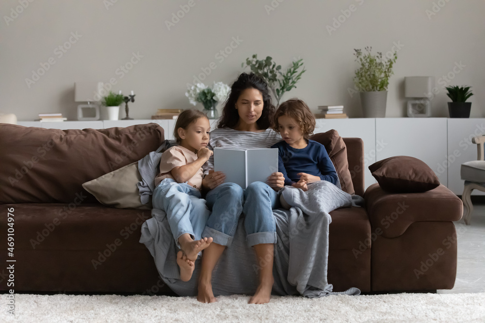 Caring mother reading fairy tale story book to kids, sitting on cozy couch at home, loving mom with little son and daughter engaged in educational activity, family spending leisure time together Caring mother reading fairy tale story book to kids, sitting on cozy couch at home, loving mom with little son and daughter engaged in educational activity, family spending leisure time together