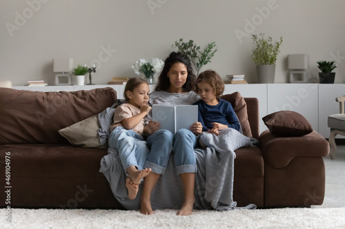 Caring mother reading fairy tale story book to kids, sitting on cozy couch at home, loving mom with little son and daughter engaged in educational activity, family spending leisure time together