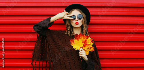 Portrait of beautiful young woman with yellow maple leaves blowing her lips sending sweet air kiss wearing a brown knitted poncho, black round hat on red background © rohappy