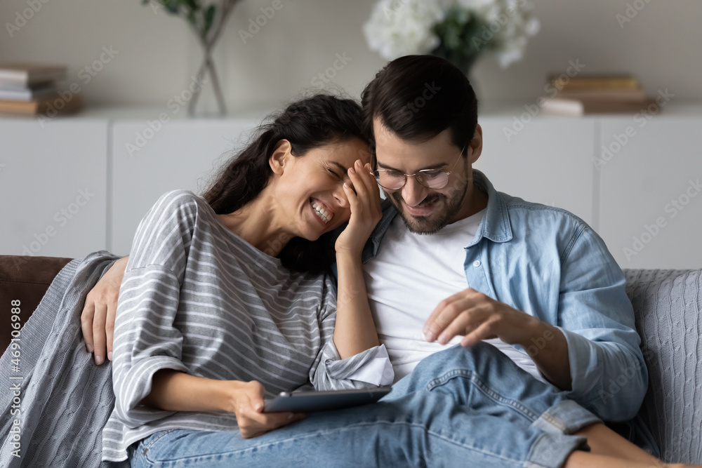 Happy man and laughing woman hugging, using tablet together, sitting on comfortable couch at home, positive couple having fun with modern device, watching funny video or movie, enjoying leisure time Happy man and laughing woman hugging, using tablet together, sitting on comfortable couch at home, positive couple having fun with modern device, watching funny video or movie, enjoying leisure time