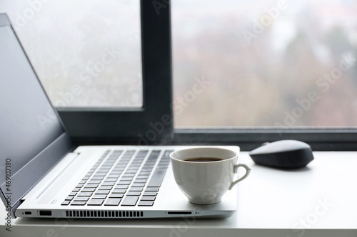 Hot coffee cup and laptop on table against the open window. Cozy workplace in home office, concept of working at morning