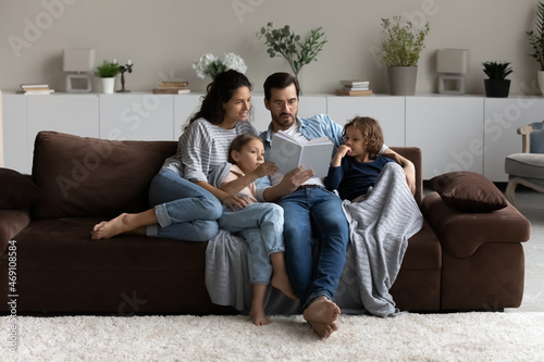 Loving parents with two little kids reading book together, hugging, sitting on cozy couch at home, smiling mother and father in glasses with adorable daughter and son engaged in educational activity