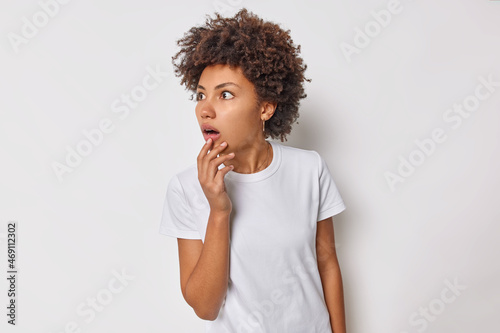 Φωτογραφία Shocked anxious woman with curly hair hears awful scary news stares concerned away cannot believe her eyes keeps jaw dropped wears casual t shirt isolated over white background