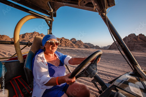 Happy elderly woman in a blue headscarf driving a buggy in the Egyptian desert with mountains in the background. © popovatetiana