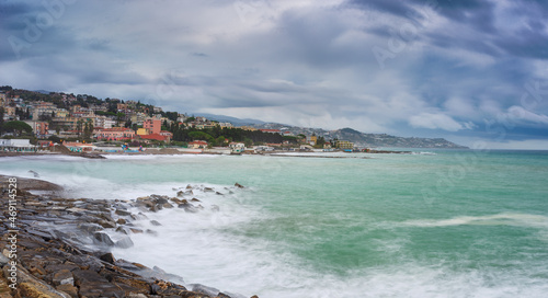Waving sea over coast line and water bay in winter, Riviera dei Fiori, Liguria, Italy. Sanremo town in the background. Dramatic stormy sky at sunset. Long exposure.