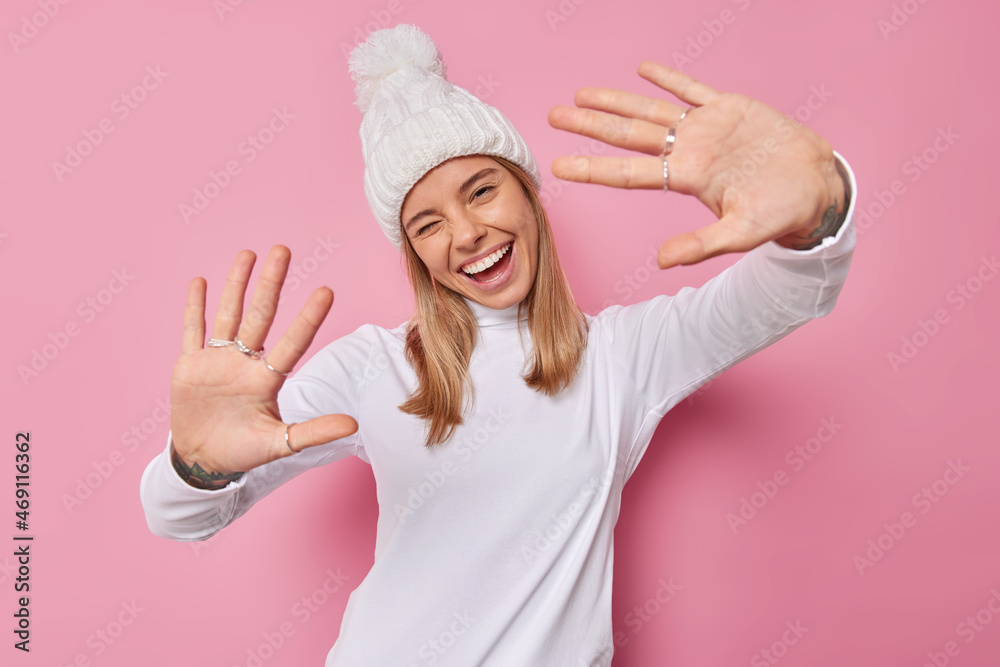 Playful cheerful woman keeps palms forward camera winks eyes and smiles gladfully wears white winter hat and turtleneck looks happily at camera isolated over pink background. Positive emotions Playful cheerful woman keeps palms forward camera winks eyes and smiles gladfully wears white winter hat and turtleneck looks happily at camera isolated over pink background. Positive emotions