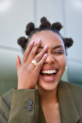 Outdoor shot of happy carefree woman smiles broadly shows white teeth makes face palm being amused by someone has bun hairstyle vivid makeup wears stylish clothes poses against blurred background