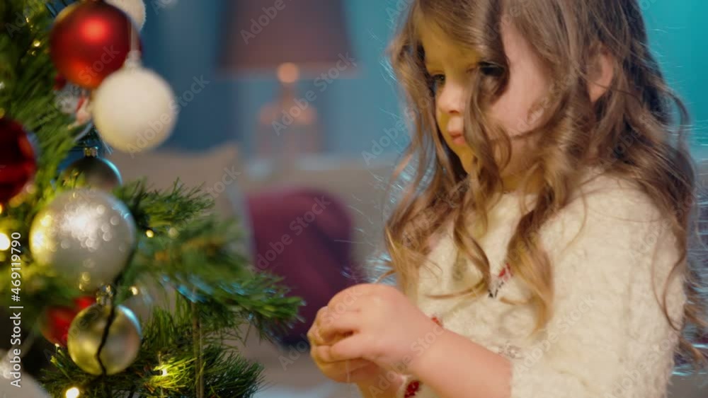 Two little girls in soft winter sweater decorating christmas tree at home with colorful toys. Happy sister creating festive atmosphere at home. Concept of family and winter holidays.