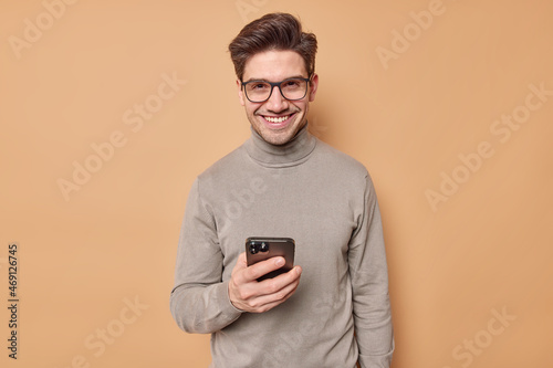Indoor shot of handsome cheerful brunet adult man uses mobile phone for chatting online wears transparent glasses and turtleneck isolated over beige background plays cool app. Technology concept © wayhome.studio 