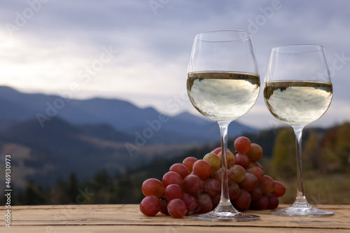 Glasses of tasty wine and grapes on wooden table against mountain landscape. Space for text