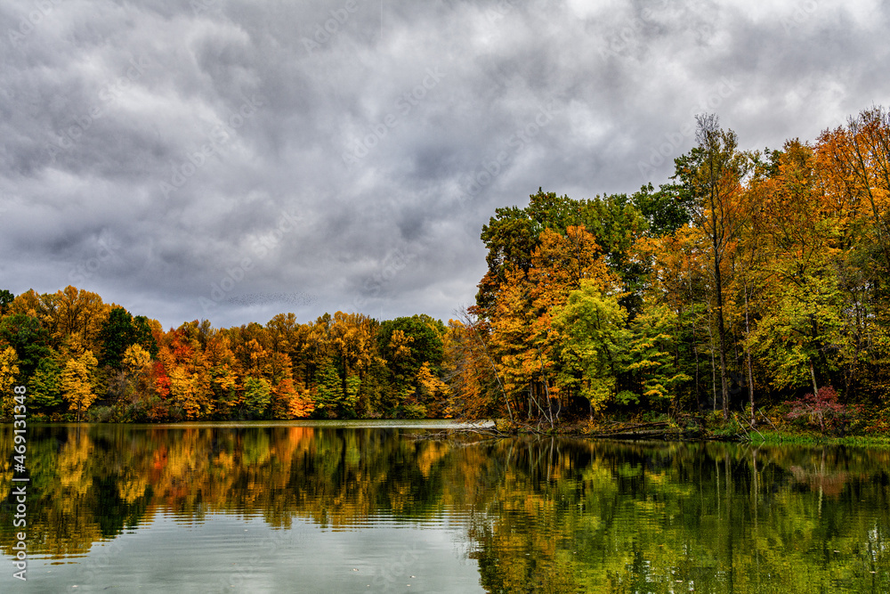 Fototapeta premium Trees ablaze with the bright colors of fall are reflected in the calm waters of Worster Lake at Potato Creek State Park in North Liberty, Indiana, as ominous storm clouds fill the sky