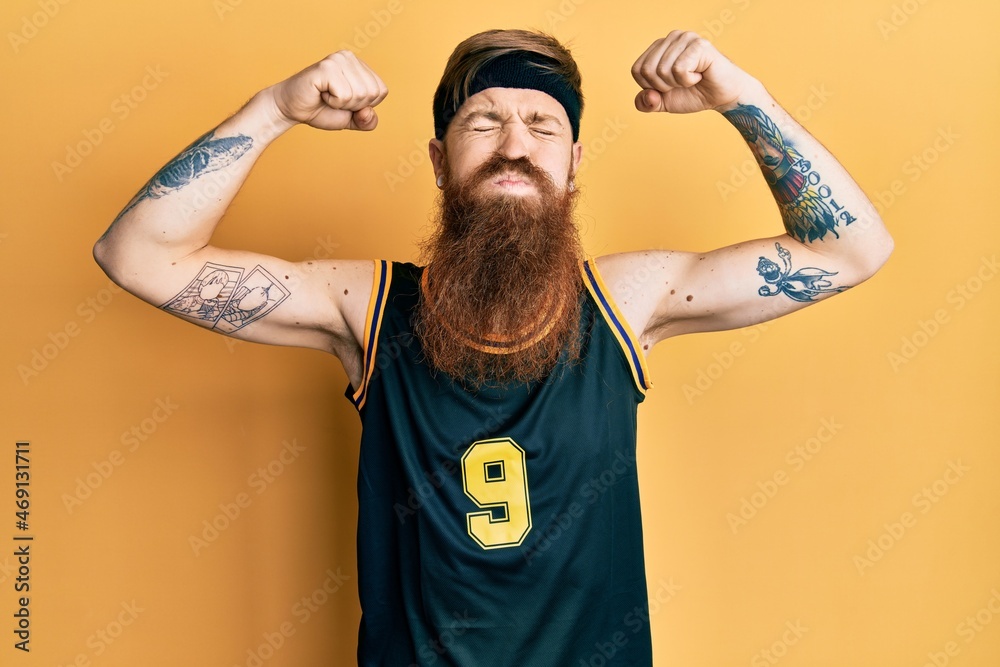 Redhead man with long beard wearing basketball uniform showing muscles puffing cheeks with funny face. mouth inflated with air, catching air.