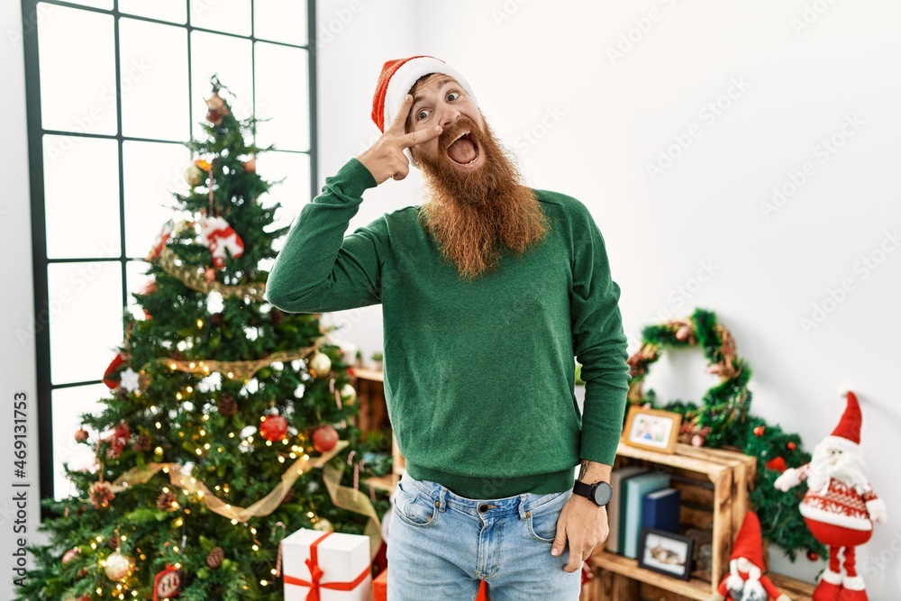 Redhead man with long beard wearing christmas hat by christmas tree doing peace symbol with fingers over face, smiling cheerful showing victory Redhead man with long beard wearing christmas hat by christmas tree doing peace symbol with fingers over face, smiling cheerful showing victory