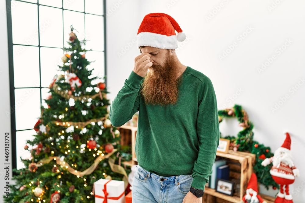 Redhead man with long beard wearing christmas hat by christmas tree tired rubbing nose and eyes feeling fatigue and headache. stress and frustration concept. Redhead man with long beard wearing christmas hat by christmas tree tired rubbing nose and eyes feeling fatigue and headache. stress and frustration concept.