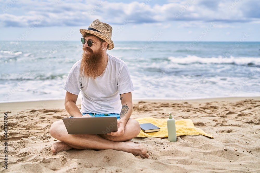 Young redhead man using laptop sitting on the towel at the beach.