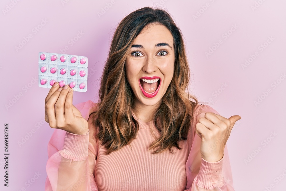 Young hispanic girl holding pills pointing thumb up to the side smiling happy with open mouth Young hispanic girl holding pills pointing thumb up to the side smiling happy with open mouth