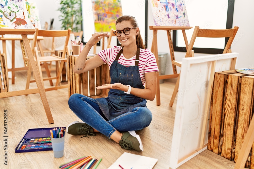 Young brunette woman at art studio sitting on the floor gesturing with hands showing big and large size sign, measure symbol. smiling looking at the camera. measuring concept.