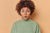 Portrait of stunned emotive beautiful woman with curly hair stares bugged eyes at camera looks concerned wears casual jumper reacts on amazing news isolated over beige background. Omg concept