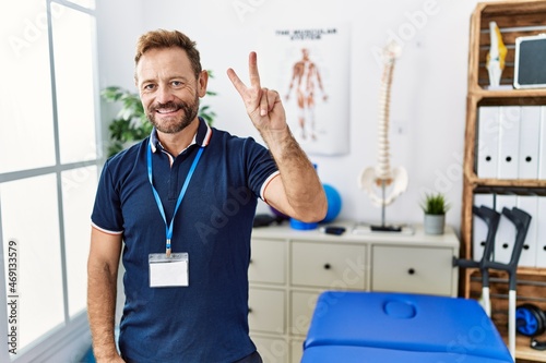 Middle age physiotherapist man working at pain recovery clinic showing and pointing up with fingers number two while smiling confident and happy.