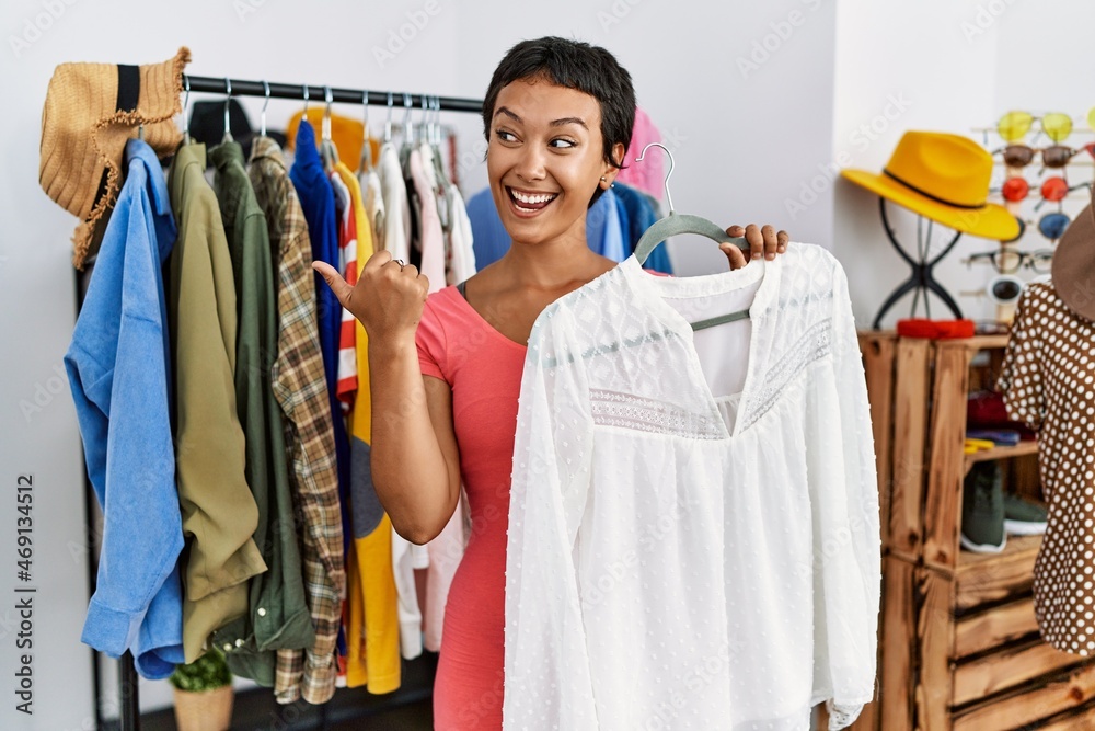 Young hispanic woman with short hair shopping at retail boutique pointing thumb up to the side smiling happy with open mouth