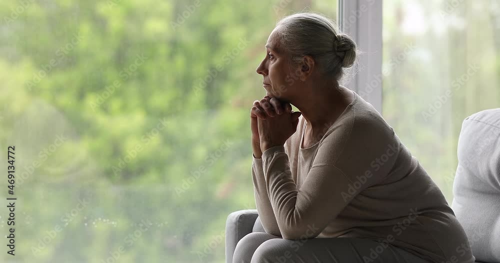 Side view of depressed older female sit on couch near picture window watch spring outside with sad look. Aged woman suffer of loneliness think of passed days of youth health problems brevity of life