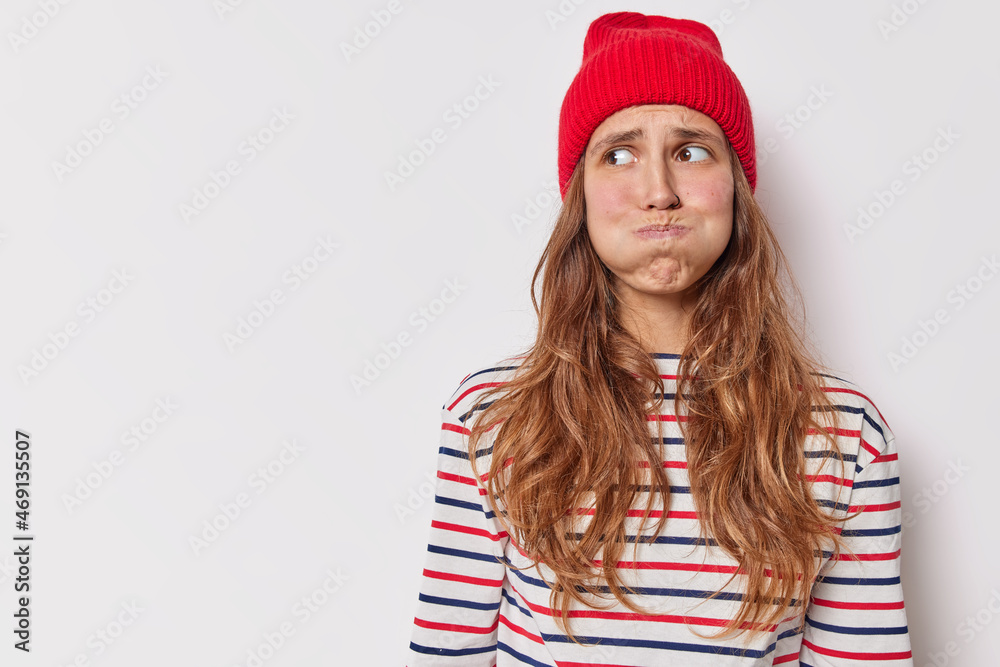 Photo of nervous young European woman blows cheeks and looks away makes grimace wears red hat casual striped jumper poses against white background with blank copy space for your information. Photo of nervous young European woman blows cheeks and looks away makes grimace wears red hat casual striped jumper poses against white background with blank copy space for your information.