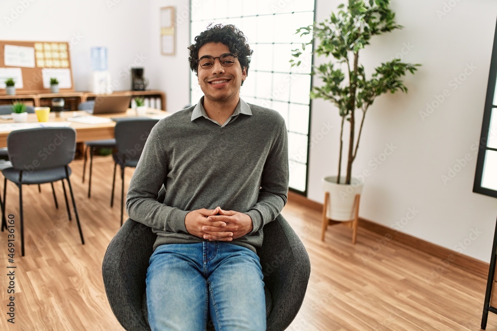 Young hispanic businessman smiling happy sitting on chair at the office.