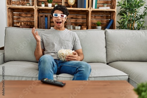Young hispanic man watching movie using 3d glasses sitting on the sofa at home.