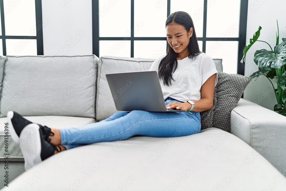 Young hispanic woman using laptop at home sitting on the sofa looking positive and happy standing and smiling with a confident smile showing teeth