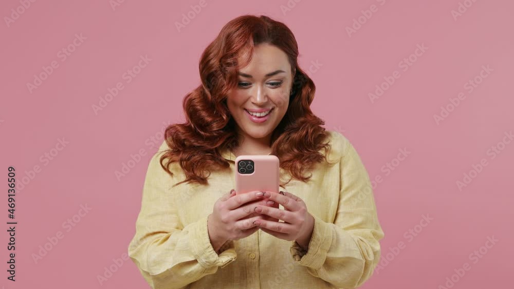 Happy charming vivid bright young ginger chubby overweight woman 20s wears yellow shirt hold using mobile cell phone typing browsing chatting send sms isolated on plain pink background studio portrait Happy charming vivid bright young ginger chubby overweight woman 20s wears yellow shirt hold using mobile cell phone typing browsing chatting send sms isolated on plain pink background studio portrait