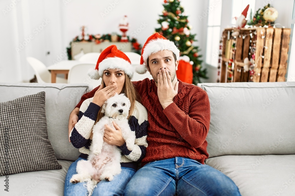Fototapeta premium Young couple of wife and husband sitting on the sofa wearing christmas hat at home covering mouth with hand, shocked and afraid for mistake. surprised expression