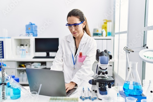 Young hispanic woman wearing scientist uniform using laptop and microscope at laboratory