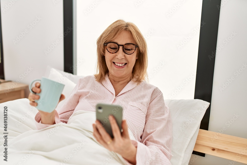 Middle age blonde woman lying on the bed using smartphone and drinking coffee at home. Middle age blonde woman lying on the bed using smartphone and drinking coffee at home.