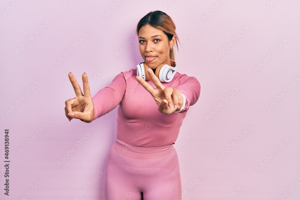 Beautiful hispanic woman wearing gym clothes and using headphones smiling with tongue out showing fingers of both hands doing victory sign. number two.