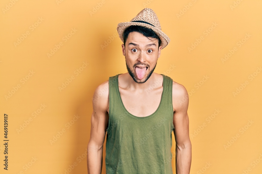 Young hispanic man wearing summer hat sticking tongue out happy with funny expression. emotion concept.