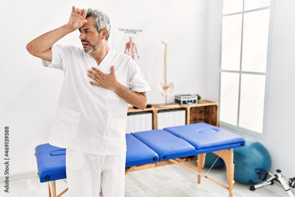 Obraz premium Middle age hispanic therapist man working at pain recovery clinic touching forehead for illness and fever, flu and cold, virus sick