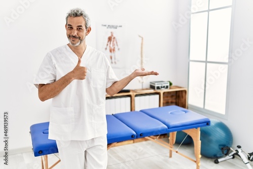 Middle age hispanic therapist man working at pain recovery clinic showing palm hand and doing ok gesture with thumbs up  smiling happy and cheerful