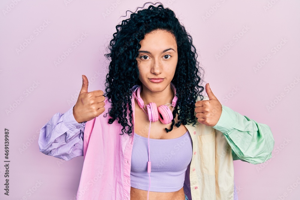 Young hispanic woman with curly hair doing thumbs up positive gesture relaxed with serious expression on face. simple and natural looking at the camera. Young hispanic woman with curly hair doing thumbs up positive gesture relaxed with serious expression on face. simple and natural looking at the camera.