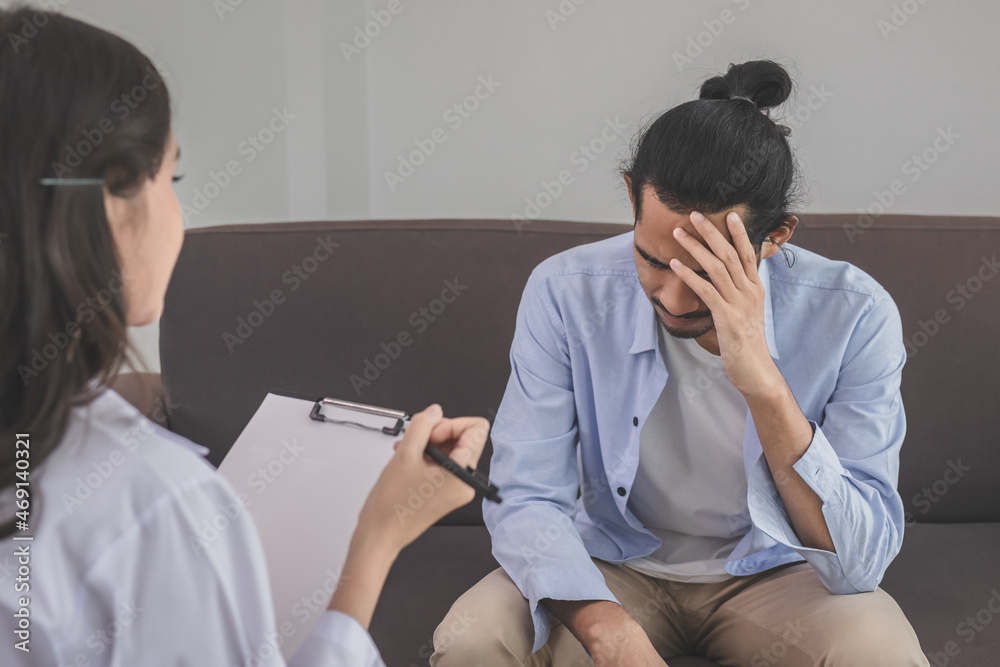 Asian young man, male has mental symptoms which must have been therapy and stress, sitting on couch to consult to psychologist during the session taking notes to find out how to treat the therapist. Asian young man, male has mental symptoms which must have been therapy and stress, sitting on couch to consult to psychologist during the session taking notes to find out how to treat the therapist.