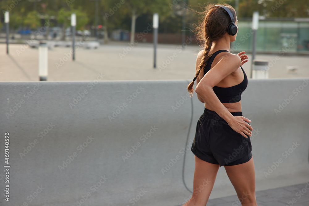Back view of motivated sportswoman in sportswear covers long distance running in urban setting keeps healthy lifestyle has regular training listens favorite music prepares for running marathon