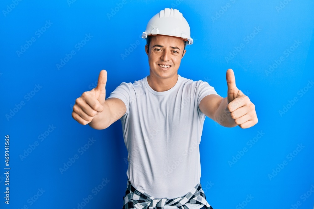 Handsome young man wearing builder uniform and hardhat approving doing positive gesture with hand, thumbs up smiling and happy for success. winner gesture.