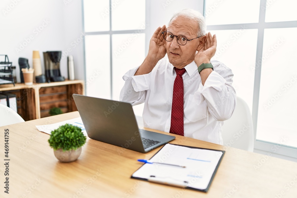 Senior man working at the office using computer laptop trying to hear both hands on ear gesture, curious for gossip. hearing problem, deaf Senior man working at the office using computer laptop trying to hear both hands on ear gesture, curious for gossip. hearing problem, deaf