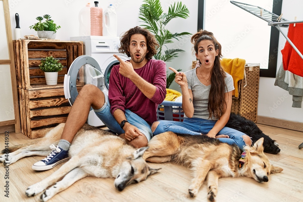 Young hispanic couple doing laundry with dogs surprised pointing with finger to the side, open mouth amazed expression.